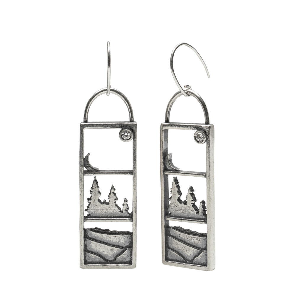 Oxidized sterling silver earrings with crescent moon, layered pine trees, and mountains, accented by a bezel-set diamond.